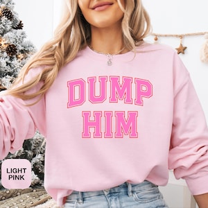 Puede incluir: Sudadera rosa claro con el texto "DUMP HIM" en una fuente rosa fuerte, de estilo universitario. La sudadera es de color rosa claro liso. Las palabras "LIGHT PINK" están en un cuadro blanco en la esquina inferior izquierda.