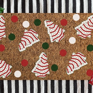 May include: A rectangular doormat with a brown cork surface features a pattern of red and white striped Christmas trees and red, green, and white circles. A sprig of holly and a wrapped gift with a red bow are in the image.
