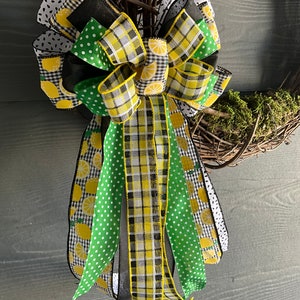 Pode incluir: Um laço verde, preto e amarelo com fita de estampa de limão. O laço é amarrado com uma fita preta e está preso a uma coroa.