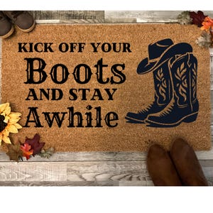 May include: A brown coir doormat with the text "Kick off your boots and stay awhile" in black lettering. A pair of black cowboy boots with a brown cowboy hat are depicted on the right side of the mat.