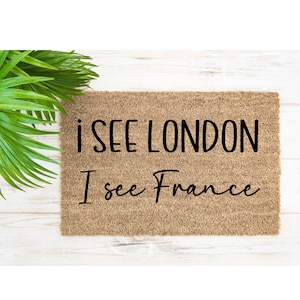 Puede incluir: Una alfombra rectangular con el texto "I SEE LONDON I see France" en escritura negra. La alfombra es de color marrón natural y está sobre una superficie de madera blanca. Se ven hojas verdes en la esquina superior izquierda.