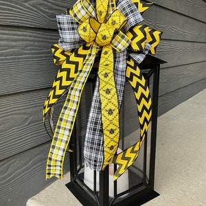 Op de afbeelding: Een zwarte lantaarn met een gele en zwarte strik erboven. De strik is gemaakt van geruit, chevron en bij-thema lint.