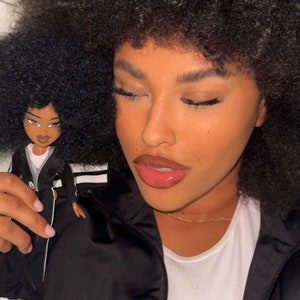 May include: A woman with a large afro hairstyle is holding a black and white doll with a similar hairstyle. The woman is wearing a black jacket with white stripes and a white shirt.