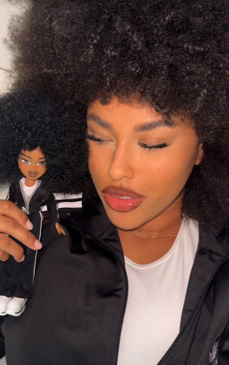 以下が含まれることがあります： 白いストライプと白いスニーカーが付いた黒いトラックスーツを着たBratz人形。人形は肌が濃く、茶色の目をし、ニュートラルな表情をしています。人形は、黒いサテンのジャケットと白いトップスを着た人のそばに持たれています。