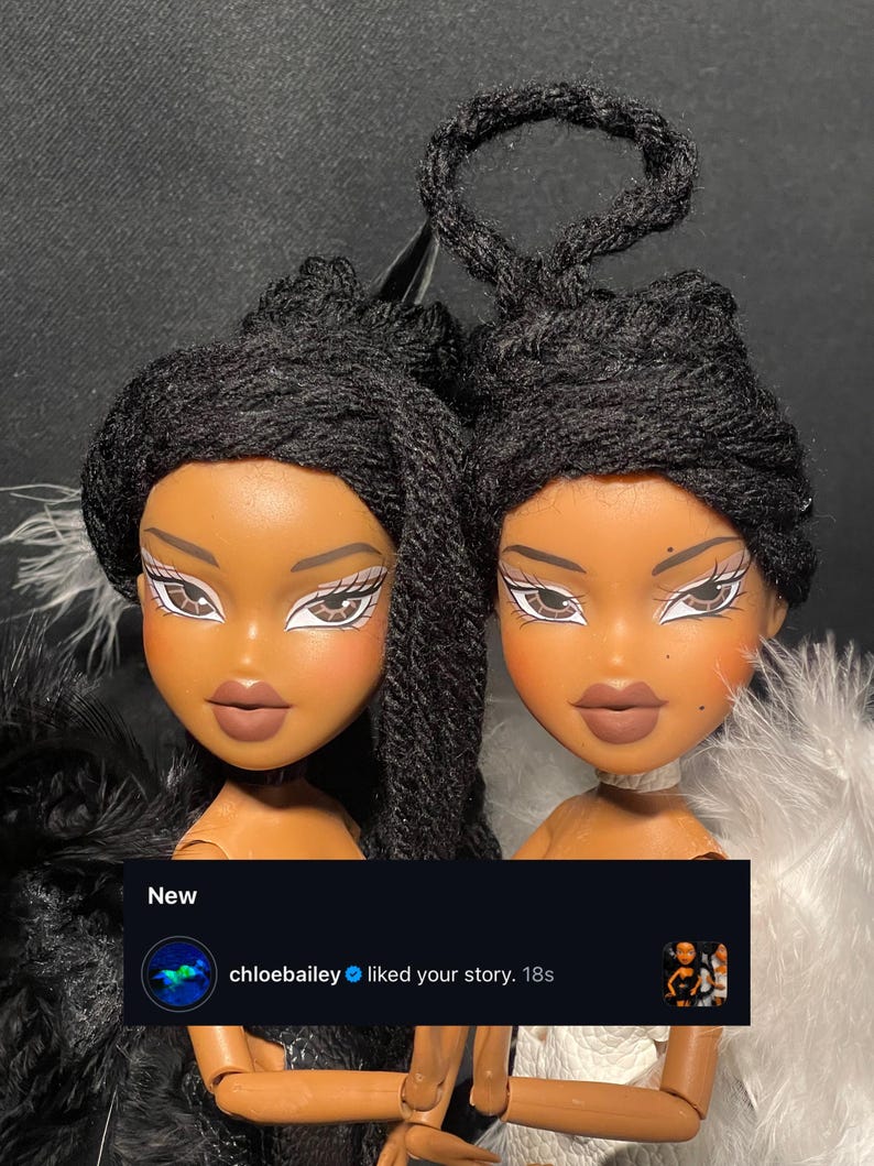 以下が含まれることがあります： 黒い三つ編みの髪と濃い肌の2体のBratz風人形。人形は、大きな目とふっくらとした唇など、詳細な顔の特徴を持っています。人形の1つは黒い羽根のボア、もう1つは白い羽根のボアです。ソーシャルメディアの通知には「New」と「chloebailey liked your story. 18s.」と表示されています。