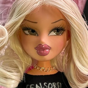 以下が含まれることがあります： ブロンドの髪の毛をしたBratz人形。黒いクロップトップを着ており、そこには「CENSORED」という文字がプリントされています。人形はふっくらとした唇、長いまつげ、金のチェーンネックレスをしています。人形の肌の色は明るいです。