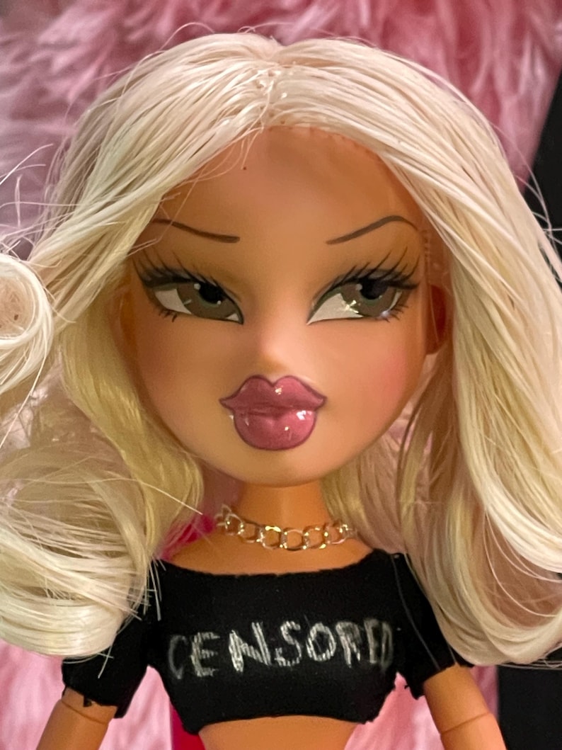 以下が含まれることがあります： ブロンドの髪の毛をしたBratz人形。黒いクロップトップを着ており、そこには「CENSORED」という文字がプリントされています。人形はふっくらとした唇、長いまつげ、金のチェーンネックレスをしています。人形の肌の色は明るいです。