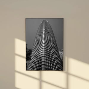May include: Black and white photograph of a tall, modern skyscraper. The building has a curved, streamlined design with a grid of windows. The perspective is from below, emphasizing the height and scale of the structure. The image is framed in a black border.