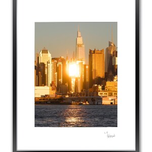 A framed photograph of the New York City skyline at sunset, with the sun reflecting brightly off the buildings and water, and a signature below the image.