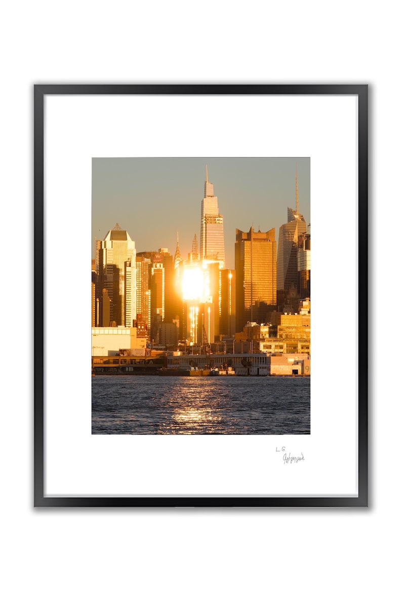 A framed photograph of the New York City skyline at sunset, with the sun reflecting brightly off the buildings and water, and a signature below the image.