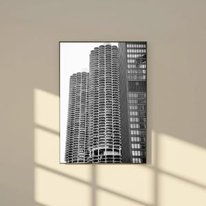 May include: Black and white photograph of a modern architectural structure. The image features two cylindrical towers with a series of rounded balconies, alongside a rectangular building with a grid of windows. The buildings are set against a bright sky.