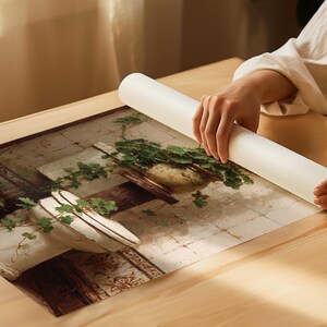 K&ouml;nnte beinhalten: Eine Person rollt einen Druck eines Gem&auml;ldes im Vintage-Stil aus. Das Kunstwerk zeigt einen wei&szlig;en Pflanztopf mit gr&uuml;nen Pflanzen und eine Steinmauer. Der Druck liegt auf einem Holztisch.