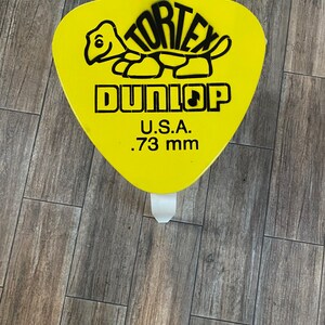 May include: A bright yellow, triangular-shaped stool featuring a black graphic of a turtle and the text TORTEX DUNLOP U.S.A. .73 mm printed on the top surface. The stool has a wide, flat seat with rounded corners and a sturdy white base structure.