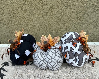 May include: Three fabric pumpkins with different patterns. The pumpkins are black with white ghost silhouettes, gray with a white geometric pattern, and gray with a white geometric pattern. The pumpkins have orange and brown stems and leaves.