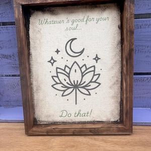 May include: A framed canvas sign with a brown wooden frame. The canvas is off-white and has a green inscription that reads "Whatever's good for your soul... Do that!"  The inscription is accompanied by a black and white illustration of a crescent moon, stars, and a lotus flower.