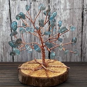 Op de afbeelding: Een decoratief boomsculptuur met een stam en takken van gedraaid koperdraad, versierd met kleine, gepolijste lichtblauwe stenen. De basis van de boom is een ronde schijf van natuurlijk hout met schors, waarop de koperdraadwortels zichtbaar zijn.