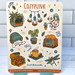 May include: A sticker sheet with the text "COZYPUNK" and various illustrations. Designs include a robot watering a plant, a cat on a radio, a mug of cocoa, and a dragonfly. The sticker sheet has a cream background.