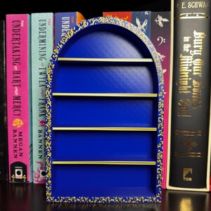 May include: A vibrant blue, arched bookshelf with four shelves, accented with gold trim and a decorative border. The bookshelf is surrounded by a collection of colorful books, creating a visually appealing display. The books have titles like "Terror at the Gates" and "Bury Our Bones".