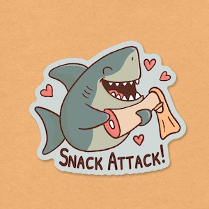 May include: A cartoon sticker featuring a smiling shark holding a piece of meat and a fin. The shark is gray and white with a wide grin. Pink hearts float around the shark, and the text "SNACK ATTACK!" is below.