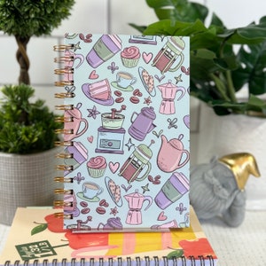 May include: A blue spiral bound notebook with a coffee and tea themed design. The cover features illustrations of coffee cups, teapots, coffee beans, and other coffee and tea related items.