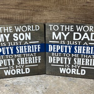 May include: Two wooden signs with the text "To the world my son/dad is just a deputy sheriff but to me that deputy sheriff is my world". The signs are painted in white and black text on a gray wood background. A blue and white thin blue line is painted across the middle of each sign.