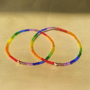 May include: Two rainbow-coloured beaded bracelets. The bracelets are made of small, colourful beads strung together. Each bracelet has a gold or silver accent bead. The bracelets are displayed on a tan surface.