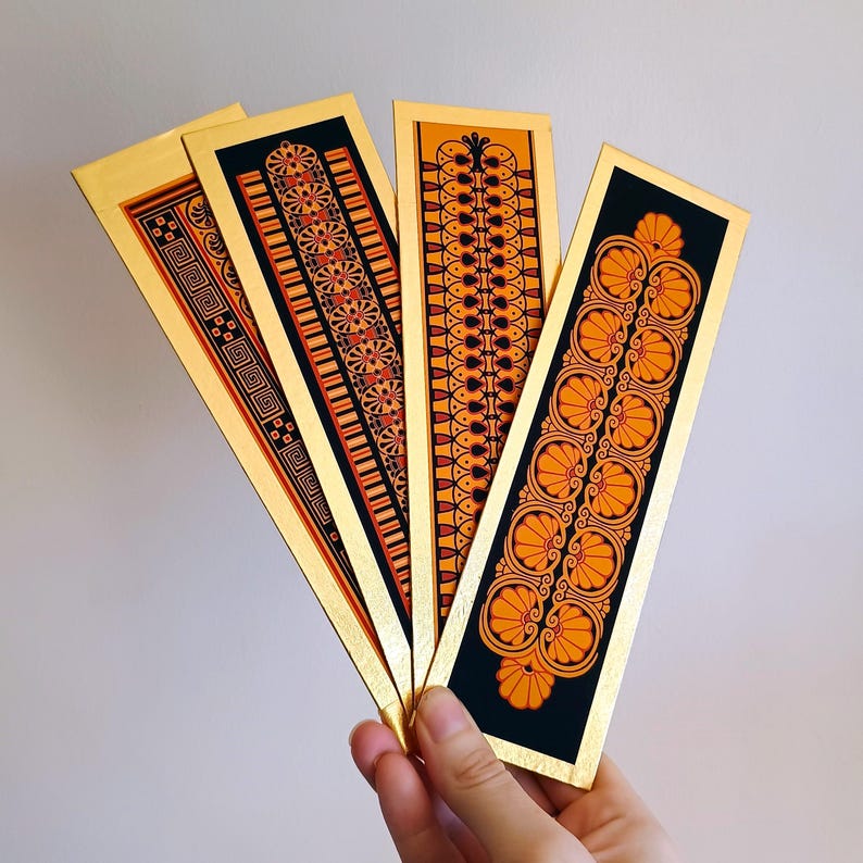 May include: Four gold bookmarks with black borders and orange and black geometric designs. The bookmarks are fanned out in a hand.