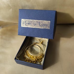 Op de afbeelding: Gouden creolen met een hangende ketting van kleine gouden kralen. De oorbellen zitten in een blauwe doos met de tekst "Yore & Lore" op de deksel.