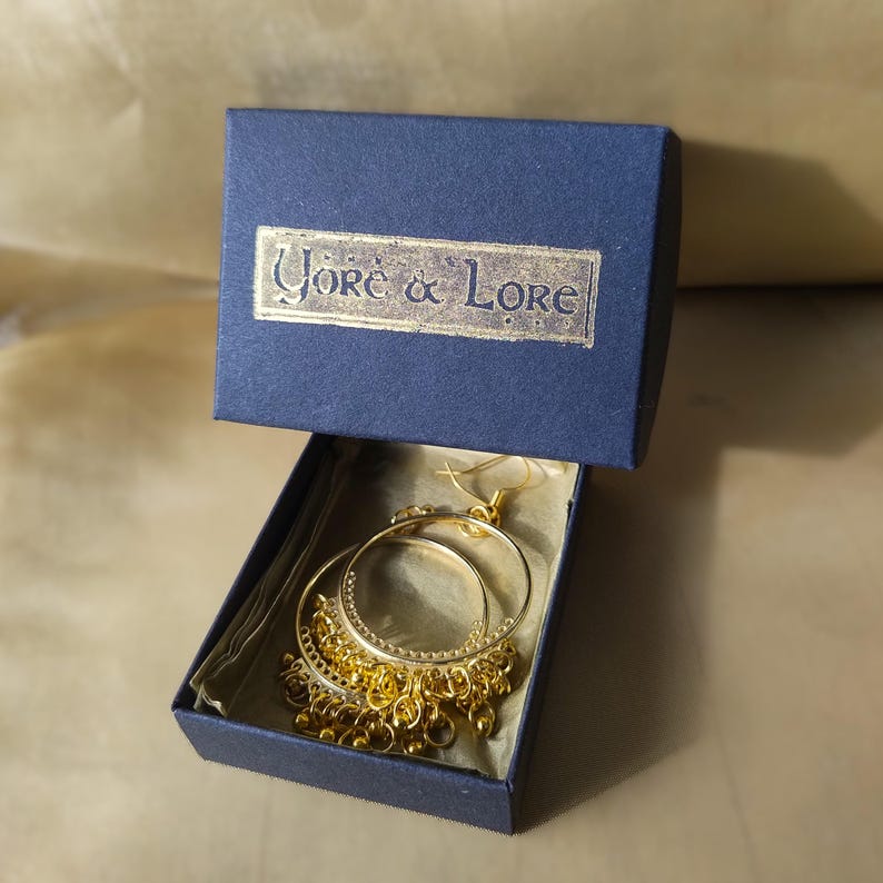 Op de afbeelding: Gouden creolen met een hangende ketting van kleine gouden kralen. De oorbellen zitten in een blauwe doos met de tekst "Yore & Lore" op de deksel.
