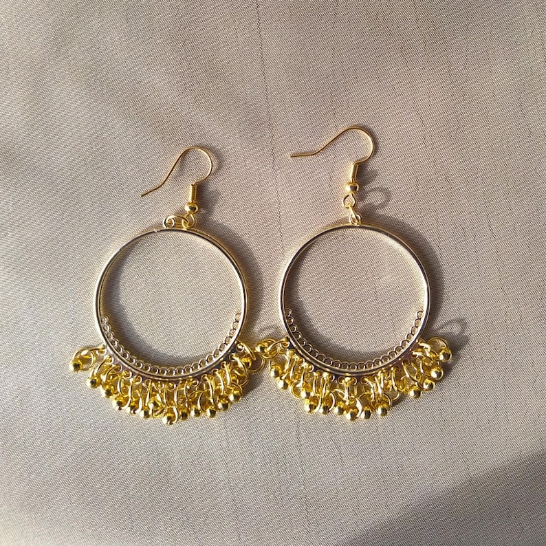 A pair of gold plated earrings in Ancient Roman style. hoops hanging from ear hooks, with many tiny gold beads hanging horizontally from the bottom of the hoops.
