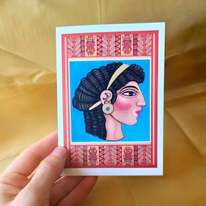 Puede incluir: Una tarjeta de felicitación con el perfil de una mujer con cabello oscuro y rizado, una diadema dorada y un pendiente dorado. La cara de la mujer es rosa con delineador negro y un labio rojo. La tarjeta tiene un fondo azul y un borde rojo y naranja.