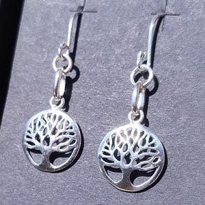 May include: A pair of silver earrings. Each earring has a circular pendant with a cut-out tree design. The earrings have a hook closure and are shown against a dark background.