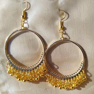 Op de afbeelding: Een paar gouden creolen met een hangende ketting van kleine gouden kralen. De oorbellen hebben een delicaat, ingewikkeld ontwerp.