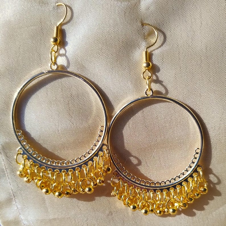 Op de afbeelding: Een paar gouden creolen met een hangende ketting van kleine gouden kralen. De oorbellen hebben een delicaat, ingewikkeld ontwerp.