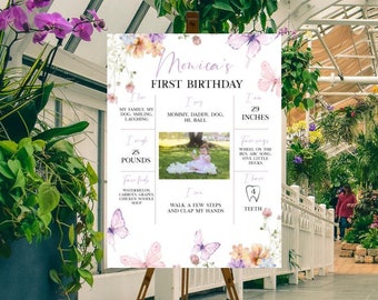 Puede incluir: Un tablero blanco con un diseño floral y mariposas. El tablero está en un caballete y tiene una foto de una niña pequeña y el texto "Amelia's First Birthday".