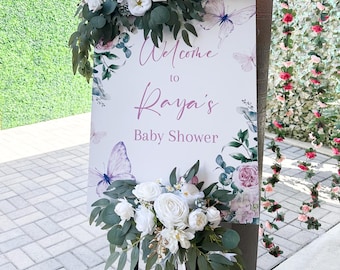 May include: A white sign with pink lettering that says "Welcome to Raya's Baby Shower" surrounded by watercolor flowers and butterflies. The sign is decorated with white flowers and greenery.