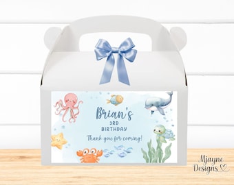 May include: A white favor box with a blue ribbon and a sticker featuring an under-the-sea theme. The sticker says "Brian's 3rd Birthday Thank you for coming!" and features an octopus, a starfish, a crab, a dolphin, a turtle, and fish.