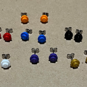 Puede incluir: Un conjunto de 10 pares de pendientes de botón con pequeñas piezas de plástico tipo LEGO en varios colores, incluyendo rojo, naranja, amarillo, azul, morado, negro, blanco y dorado.