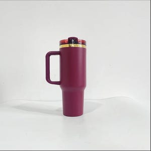 May include: A burgundy stainless steel travel mug with a gold lid and a handle. The mug is insulated and has a double-wall construction.