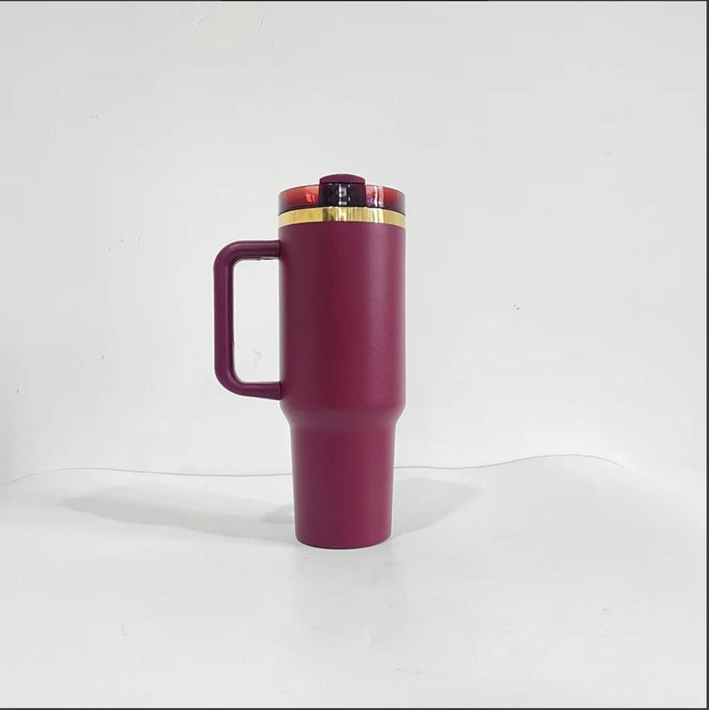 May include: A burgundy stainless steel travel mug with a gold lid and a handle. The mug is insulated and has a double-wall construction.