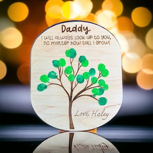 May include: Wooden plaque with a tree design created with green fingerprints. The plaque has the text "Daddy" and "I will always look up to you, no matter how tall I grow!" with "Love, Haley" at the bottom. The plaque is light brown.