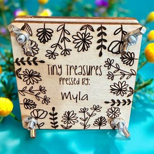 May include: A wooden box with floral and leaf designs, secured with metal hardware. The text "Tiny Treasures Pressed By: Myla" is centered on the front. The box is surrounded by yellow and purple flowers against a turquoise background.