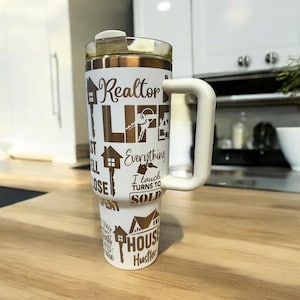 May include: White stainless steel tumbler with a brown handle and gold lettering. The tumbler has a variety of real estate-themed phrases printed on it, including "Realtor Life", "Everything I Touch Turns to Sold", and "House Hustler".