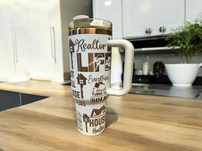 May include: White stainless steel tumbler with a brown handle and gold lettering. The tumbler has a variety of real estate-themed phrases printed on it, including "Realtor Life", "Everything I Touch Turns to Sold", and "House Hustler".