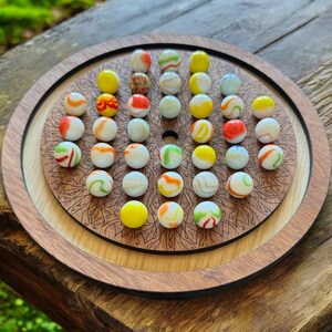 May include: A wooden game board with a circular design and a mandala pattern. The board is filled with colorful marbles in shades of white, red, yellow, and green.