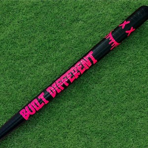 Puede incluir: Bate de b&eacute;isbol negro con las palabras "BUILT DIFFERENT" en letras de color rosa brillante. El bate tambi&eacute;n tiene detalles en rosa y dos marcas "XX" rosas cerca de la parte superior. El bate est&aacute; sobre un fondo verde.