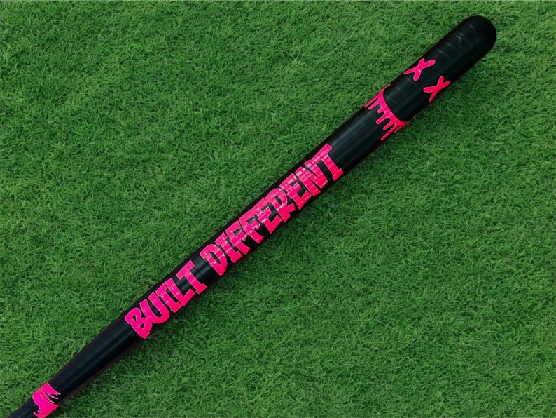 Puede incluir: Bate de b&eacute;isbol negro con las palabras "BUILT DIFFERENT" en letras de color rosa brillante. El bate tambi&eacute;n tiene detalles en rosa y dos marcas "XX" rosas cerca de la parte superior. El bate est&aacute; sobre un fondo verde.