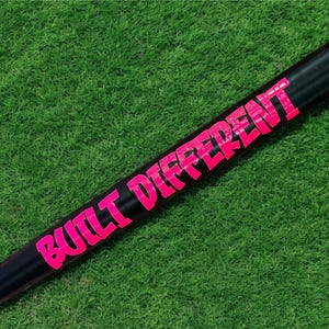 Puede incluir: Un bate de b&eacute;isbol negro con las palabras "BUILT DIFFERENT" en letras de color rosa brillante. El bate se encuentra sobre un fondo de hierba verde.