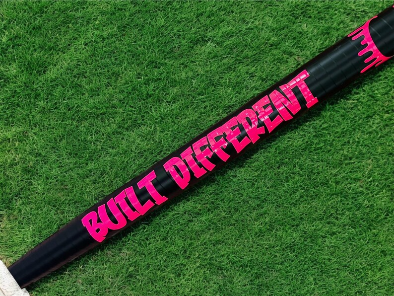 Puede incluir: Un bate de b&eacute;isbol negro con las palabras "BUILT DIFFERENT" en letras de color rosa brillante. El bate se encuentra sobre un fondo de hierba verde.