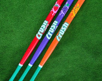 May include: Three colorful baseball bats with the "Crush" logo. The bats are red, purple, and orange, each with a different fruit graphic: strawberry, grapes, and a citrus slice. The handles are green with yellow ends.